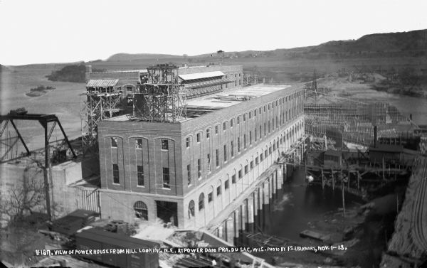 Elevated view from the hill, looking northeast at the power house at the Prairie du Sac dam. Much of the brickwork has been completed. Concrete forms for the lock are apparent on the right. The narrow gauge railroad is also visible on the right.