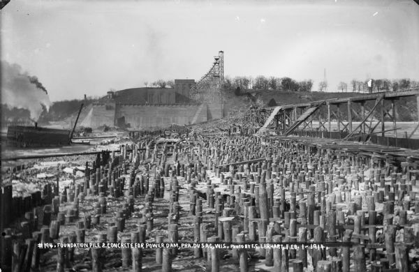 A view looking west toward the lock and power house at the Prairie du Sac dam. Pilings and the elevated trestle are in the foreground. Men can be seen working behind the pilings in the background.