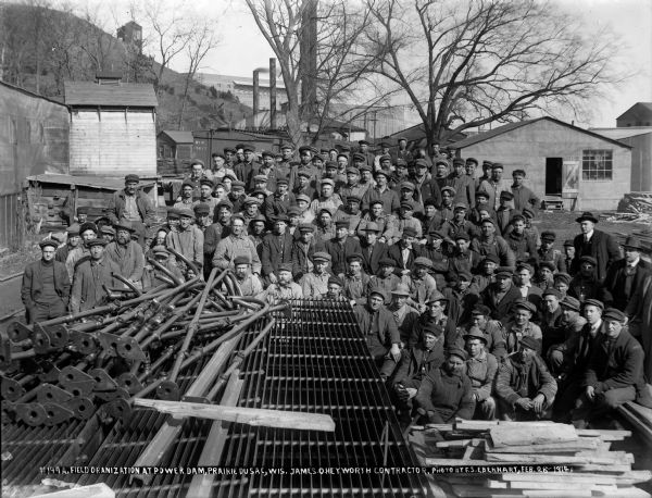 Group portrait of workers assembled with construction materials south of the power dam at Prairie du Sac. Three men in suits are on the right. The smokestacks of the temporary power plant are behind the group, with the new power house in the background.