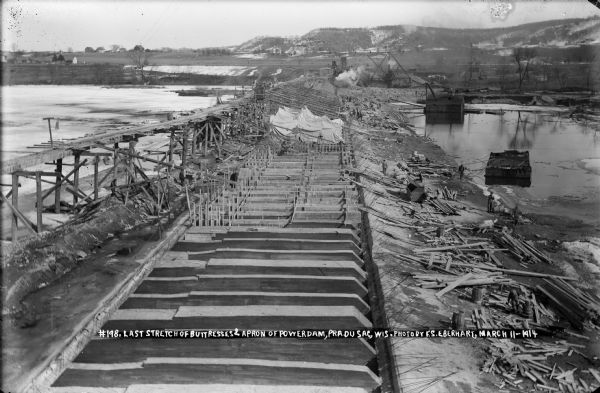 Elevated view looking east from the lock at the Prairie du Sac dam. Recently completed buttresses are in the foreground, with earlier stages of construction visible, including concrete forms. The concrete chutes extend from the elevated trestle, and the concrete plant is visible on the east bank. Note that the flow of the river has been diverted through the completed lock and power house. Men are working on the site.