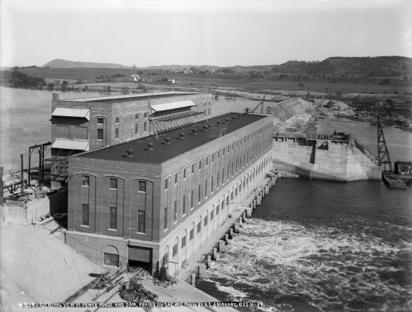 Elevated view looking east across the power house, lock, and uncompleted portion of the dam. The concrete plant is seen on the far bank. The rising water of Lake Wisconsin upstream from the dam is submerging trees. Workmen are standing on the wall of the lock.