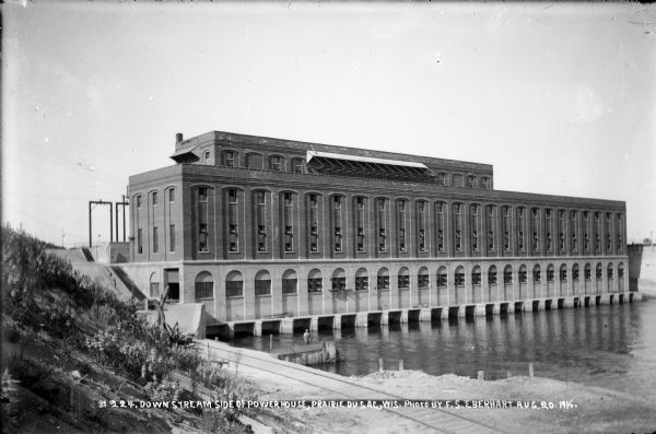 View of the south, or downstream, side of the power house at the Prairie du Sac dam. A man is standing on pilings near the bank; the railroad extension is in the foreground.