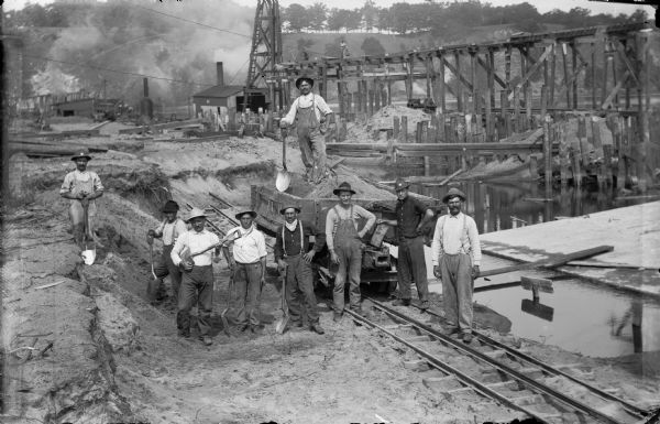 Elevated view of a group of workers posing on the east bank of the Wisconsin River at the power dam construction site. One man is strumming his shovel like a banjo; another man is standing atop a railroad car. They are working on the track for the narrow gauge electric railroad. The high trestle and steam-powered pile driver are in the background.