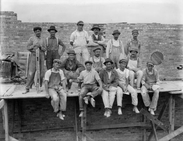 A group of bricklayers posing with the tools of their trade in front of an unfinished brick wall at the Prairie du Sac dam site. Note the wooden form for a circular opening in the wall.