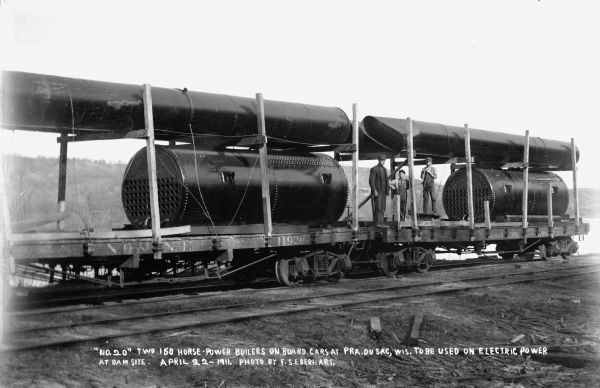 Two men and a boy are posing on rail cars carrying two 150 horse-power boilers for use in the temporary power plant at the dam construction site at Prairie du Sac. The cars also carry sections of smokestack.