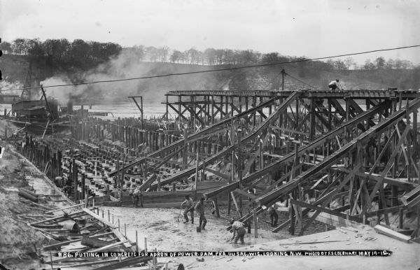 Elevated view of workers using shovels to level fresh concrete at the base of the chutes from the high trestle. The steam powered pile driver is at work in the background.