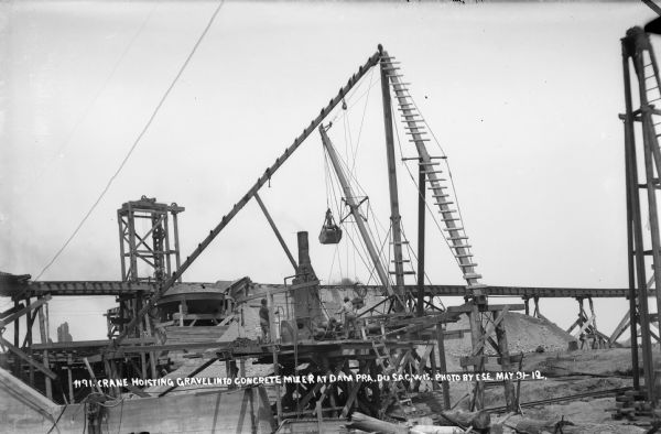 Workers maneuvering a load of gravel into a concrete mixer at the plant on the east bank of the river. The east abutment is in the foreground, left.