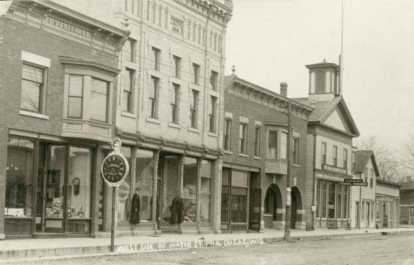 Photographer's jewelry store and clock repair shop with a watch-shaped sign in front. In the window are a clock, jewelry box, and a rack with paper goods, possibly photographic postcards. The next building, with the inscription, "J. Hatz 1889," housed a dry goods and grocery store. The sign on the two-story wooden building reads "F.O. Leigh General Hardware." Caption reads: "West Side of Water Street, Pra. du Sac, Wis."