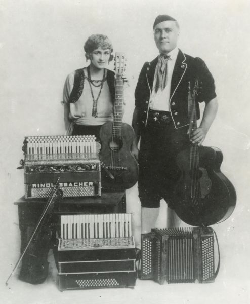 Portrait of Iva and Otto Rindlisbacher standing with various musical instruments. Otto is in Swiss costume.