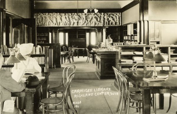 Interior view of the Carnegie Library, Richland Center, WI. Two people are seated at a table reading at the far left. A frieze decorates the wall above a doorway between rooms.