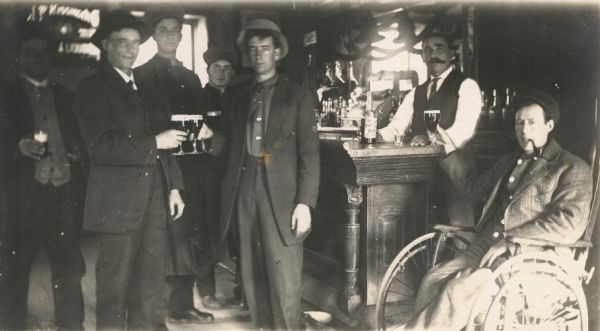 Group of men in a tavern making a toast. The bartender is behind the bar and the man at the right is in a wheelchair and smoking a pipe.