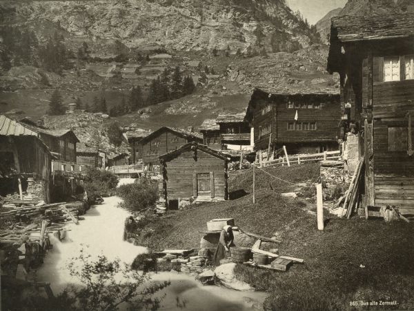 Houses by a stream in Zermatt, Switzerland. A woman is doing laundry (?) in the foreground.