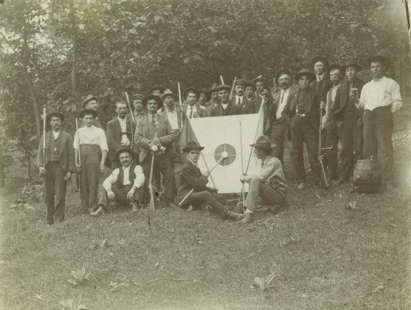 A Swiss "Schutenfest" club, affiliated with the Central Shooting Society of Wisconsin, started about 1877 and lasted until about 1920. Group of men posing with guns and a target. A man on the right has a barrel at his feet and holds up a glass of beer.