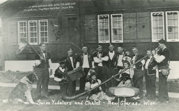 Group of people in Swiss costume with large wheel of Swiss cheese, alpine horn and other instruments, and a St. Bernard in front of the Chalet of the Golden Fleece.Identified left to right: Paul Grossenbacher, unknown, Herman Matzinger, Dorthea Widmer (seated), John Amacher, Oswald Scheider, Karl Mueller, Casper Yaun, Anna Leuthi (seated), Ernst Thierstein, and Rudy Burkhalter.