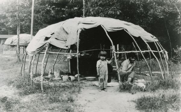 A Winnebago (Ho-Chunk) man and child inside a wigwam with the sides rolled up. A cat is walking in the grass in front of the man.