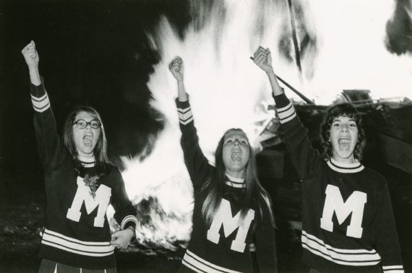 Three cheerleaders from Mayville High School perform in front of a bonfire.