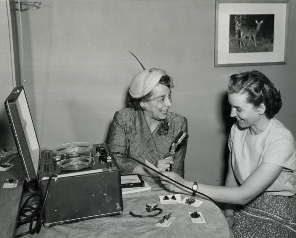 Aline W. Hazard (in hat) of WHA Radio, recording and interviewing Mrs. Robert Jahnke (Corrine) at Forest Acres Deer Farm. Mrs. Jahnke holds a pair of earrings that she made from acorns and pine cones.  These were sold at their gift shop for the deer farm.  The deer farm was started in 1955, and was located adjacent to their home, 3 1/2 miles south of Cornell on Hwy 27. There are other earrings and a watch on the table in front of the women.