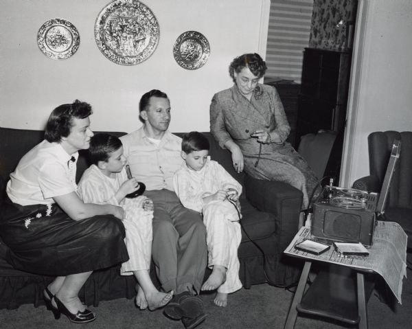 Aline W. Hazard checks a stopwatch as a boy dressed in pajamas speaks into a microphone. The boy is seated on a couch with his parents and younger brother. A reel to reel recorder sits on a table nearby.