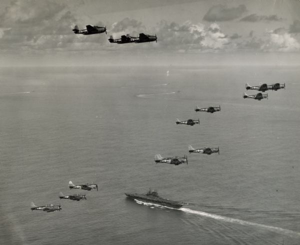 Aerial view of three TBF's (all above the horizon) and eleven SBD's, which participated in Pacific Fleet's carrier-strike against Japan's bases in the Palau Islands, off for the attack. Warships, including an aircraft carrier, are visible in the ocean below.