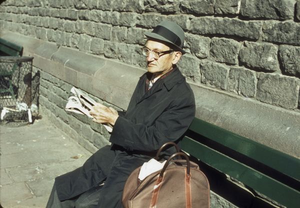 An elderly man sitting on a bench reading a newspaper near the World Trade Center.