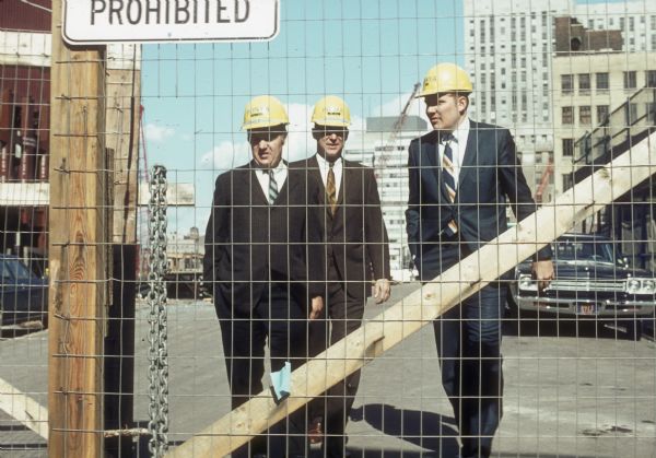 Three men in hard hats, possibly architects, walking behind a fence at the World Trade Center construction site.