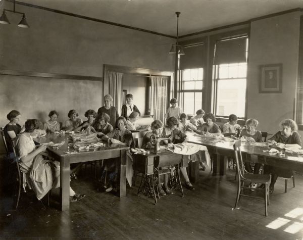 Young girls at work in the Rochester Short Course sewing laboratory, with Nellie Kedzie Jones supervising their work. Many girls are doing handwork and some are using machines. The young woman in the foreground is using a Singer sewing machine.