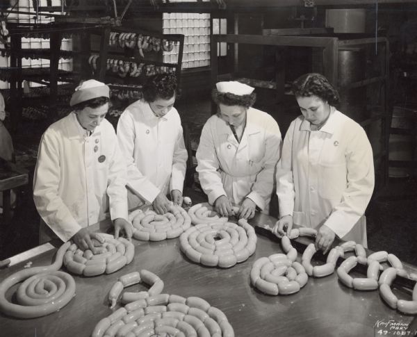 Four women butcher workers in white lab coats hand tie sausage links at the Oscar Mayer plant.