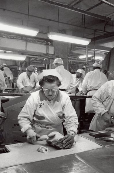 A woman worker on the poultry line at an unidentified packer, probably in Chicago. All of the other workers, all of whom were represented by the Amalgamated Meatcutters and Butcher Workmen of North America, are men. The peace symbol of the 1960s has been drawn on the woman's uniform.