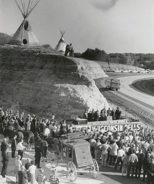 Dedication ceremony for a section of Interstate Highway I-90 near the Wisconsin Dells, with a group of Native Americans watching from above. A Dells Fargo Express Wagon is parked near the speakers' stand.