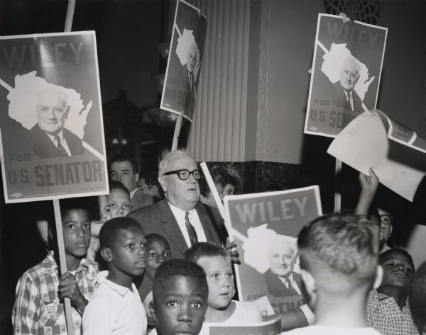 Senator Alexander Wiley, in the lobby of the Schroeder Hotel in Milwaukee, with a group from the Boys Club holding campaign signs.  They presented him with an honorary membership. The senator was defeated in his attempt for re-election by Gaylord Nelson.