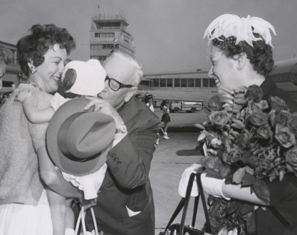 At Mitchell Airport, Senator Alexander Wiley greets the young daughter of Mr. and Mrs. Underborg of Waukesha with a kiss. Mrs. Wiley, holding a bouquet of roses, looks on. Wiley was campaigning for re-election.