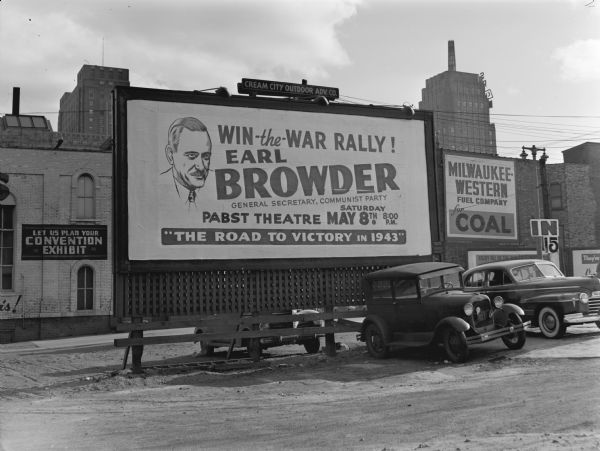 A Milwaukee billboard advertising a lecture by Earl Browder, general secretary of the Communist Party-USA. The sign reads "Win the War Rally! Earl Browder, general secretary, Communist Party" and "The Road to Victory in 1943." Two cars are parked next to the parking lot billboard and industrial buildings rise in the background.