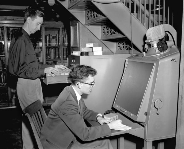 Kenneth Duckett and Alan E. Kent, historians employed by the State Historical Society of Wisconsin, using the microfilm reader in the Society's old, first floor manuscript room.  Bound document collections can be seen in the closed shelving behind them.