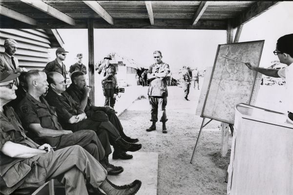 Secretary of Defense Melvin Laird, and several cigar-smoking officers are listening to a presentation about the war in Vietnam.