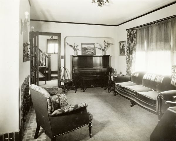 Living Room of the 1928 demonstration home in Kohler, Wisconsin, showing health carpet, tan murescoed wall, fireplace with wrought iron fire set, della robbia with dull blue background, brown and old rose tapestry fireside chair, stool and piano. Also visible is the entryway showing a desk, an oil painting from the Chicago Galleries Association, green gauze curtains that match those in the living room. This photograph was included in a scrapbook presented to Marie C. Kohler, Chair of the better homes project.