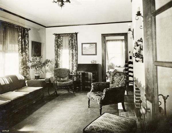 Living Room, looking toward the sun room, showing the green velvet sofa, coffee table with fuschia plant, desk, over curtains (tan background, with old rose and purple flowers), wrought iron curtain rods, oil paintings from the Chicago Galleries Association, mulberry tapestry arm chair, and ladder back chair.  This photograph was part of a scrapbook presented to Marie C. Kohler, chair of the annual demonstration home project.