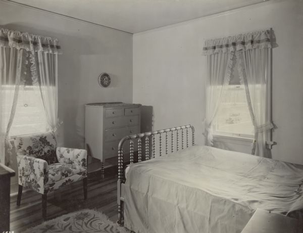 Bedroom in the 1928 Demonstration Home in Kohler, Wisconsin. The decor features jade green furniture, a flowered chair, and corn-colored curtains banded with green stripes and pink roses. The bed is covered in an old gold rayon taffeta spread with appliqued flowers. There is a della robbia plaque on the wall. This photograph was part of a scrapbook presented to Marie C. Kohler, who was the chair of the annual event.