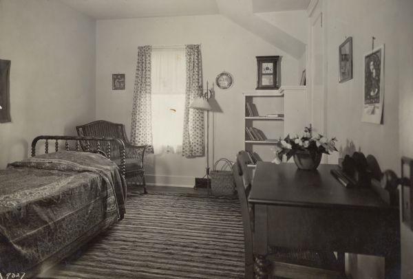 Bedroom in the 1929 Demonstration Home in Kohler, Wisconsin, decorated for a young boy.  The spool bed is covered with a green, yellow and red India coverlet.  The curtains are colorful.  There is an old-fashioned clock, a desk, a highboy, a wicker chair, and a built-in bookcase.  The rug is a handwoven rag rug of black, blue, brown, yellow and green.  The all wood floor is stained dark.
