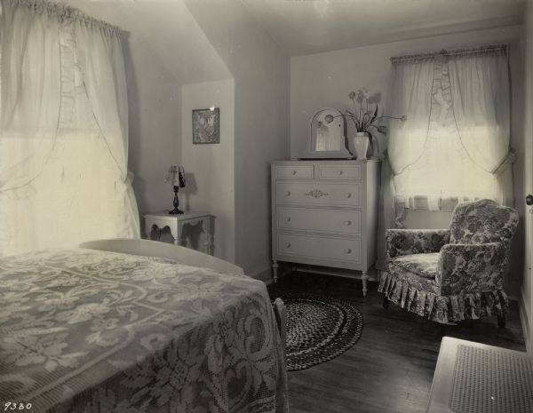Guest room in the 1929 demonstration home in Kohler, Wisconsin. The decor featured lavender organdie curtains, a flowered cretonne covered chair, a cane seat bench and a highboy with painted floral designs. The bedspread is Chinese filet, green over lavender sateen. The rugs are braided in black, green and yellow. The walls are light cream moresco. This photograph was part of a scrapbook presented to Marie C. Kohler, chair of the event.