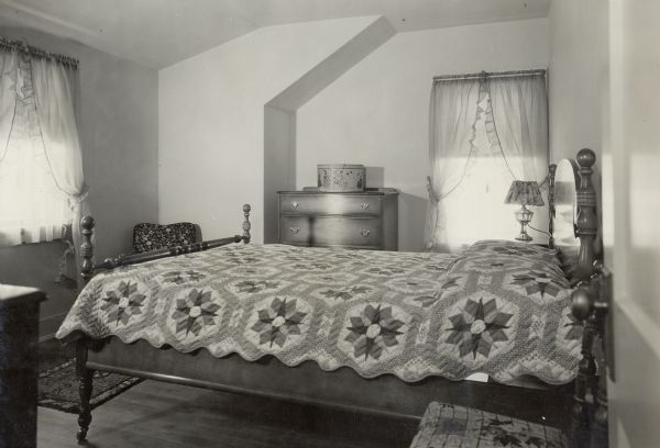 Bedroom in 1929 Demonstration Home in Kohler, Wisconsin. The bedroom is painted in buff moresco.  There is a braided rug and a hooked rug on the floor.  It is furnished with a walnut set, and a cretonne chair in greens, yellows, and blacks. The curtains are green organdie,  The bedspread is a lavender print patchwork quilt, valued at $4.50. There is also a green painted chest with vari-colored floral designs. This photograph is part of a scrapbook presented to Marie C. Kohler, chair of the annual event.