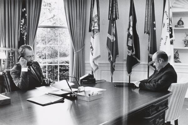 President Richard Nixon speaking on the telephone in the Oval Office of the White House, while Secretary of Defense Melvin Laird is waiting to report on his recent trip to the Far East.
