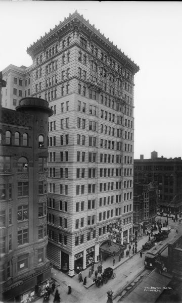 Elevated view of Majestic Theater Vaudeville building, with pedestrians on the street, along with a streetcar and horse-drawn vehicles. The Schlitz Hotel is on the right, on the south side of West Wisconsin Avenue between North 2nd and North 3rd Streets.