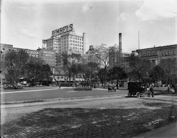 Park near hotels and Gimbels building. Caption reads: "View N.E. from C. M. & St. P. Depot". Presently, it is Zeidler Union Square on W. Everett Street between N. 2nd and N. 3rd streets. This view is seen from the front of the Milwaukee Road Depot, with the Public Service Building (now We Energies) on the right.