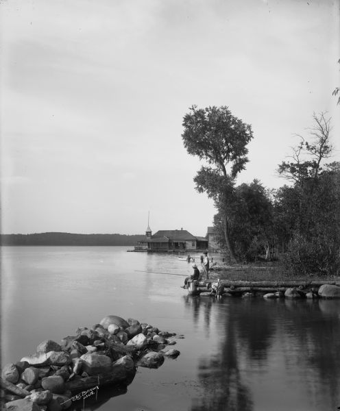 Three boys on the shore of Pewaukee Lake. One boy is fishing, while the other two observe. A boat dock and boathouse are in the background.
