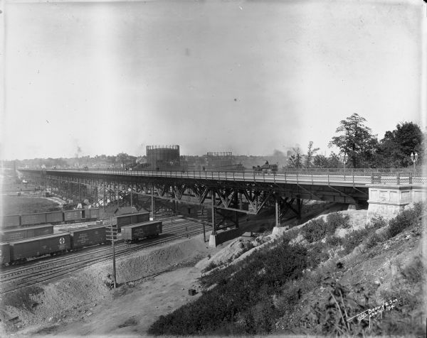 27th Street viaduct looking north, Menomonee Valley area, with gas holders of the Milwaukee Gas Light Co. in the background. Pedestrians and wagons are crossing the bridge. Railroad tracks are under the viaduct.