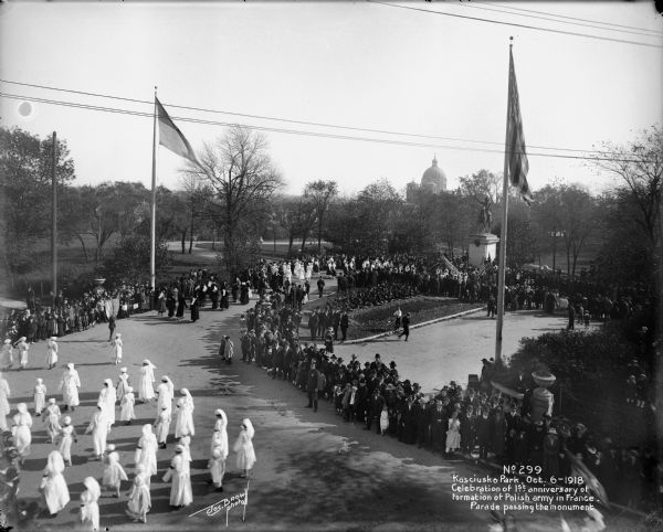 Elevated view of Polish parade in Kosciuszko Park at W. Becher and S. 8th Streets, with St. Josaphat's Basilica in the background. The Kosciuszko statue was later moved to the Lincoln Avenue side. Caption on glass plate reads: "Celebration of first anniversary of formation of Polish army in France. Parade passing the monument."