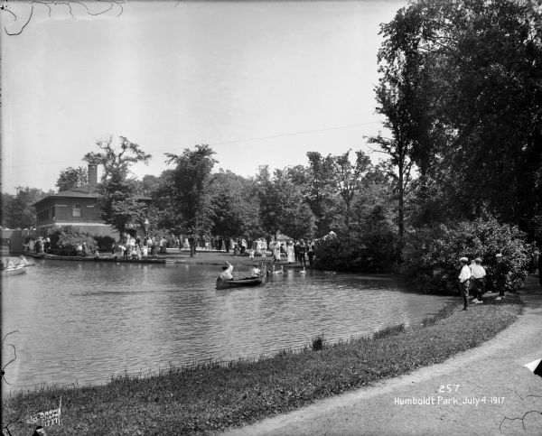 Children canoeing in the pond with swans, while other children are watching. There are adults and children in the background near a building.