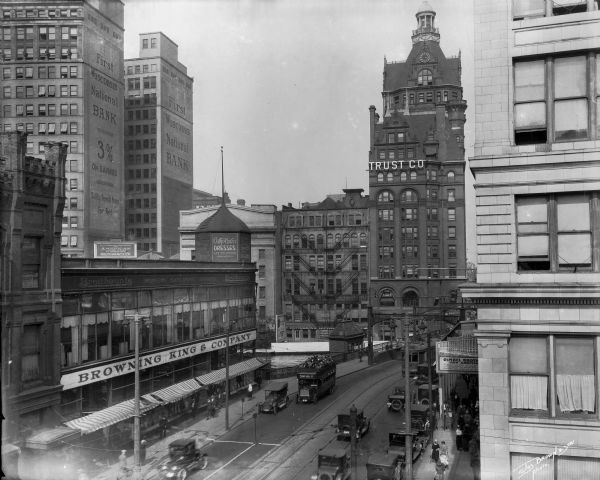 Elevated view east from N. Plankinton Avenue, W. Wisconsin Avenue is bustling with automobiles, pedestrians, a cable car, and a double-decker bus. Landmarks right to left: Gimbels Department Store, Pabst Building (ornate tower), The First Wisconsin National Bank and Browning King (men's clothing) in the foreground.