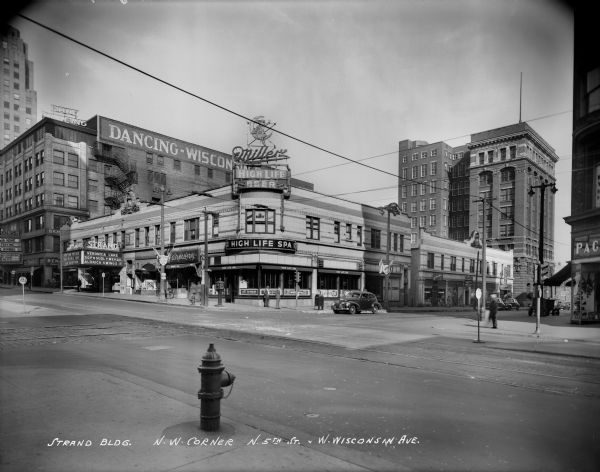 The corner of N. 5th Street and W. Wisconsin Avenue. Landmarks from left to right: Wisconsin Roof (dancing), Strand Theater, Miller High Life Spa, and the Wisconsin Telephone (the eight-story building). "So Proudly We Hail!," a 1943 film starring Veronica Lake, is posted on the Strand Theater's marquee.