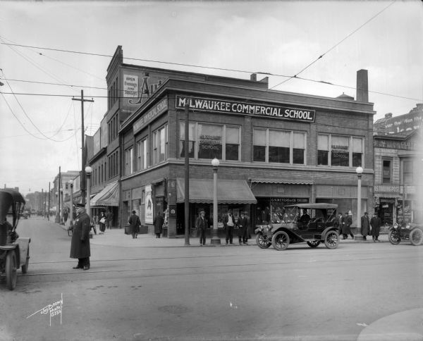 Northwest corner of 3rd and Wells Street, facing the Milwaukee Commercial School. A police officer directs traffic in the foreground, with pedestrians on the sidewalk in background.